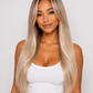 A woman with long, straight, light blonde hair with dark roots, wearing a white tank top and light blue jeans, photographed against a neutral light gray background.