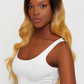 A woman modeling a long wavy wig with a dark brown to golden blonde ombre transition, paired with a white tank top and beige pants, against a plain light gray background.