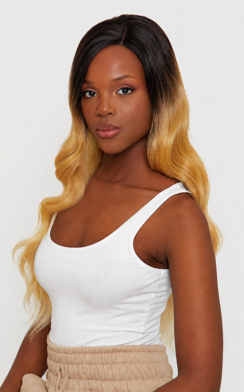 A woman modeling a long wavy wig with a dark brown to golden blonde ombre transition, paired with a white tank top and beige pants, against a plain light gray background.