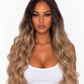 A woman product a long, wavy wig with dark brown roots and warm blonde ombré highlights, wearing a white tank top against a plain white background.