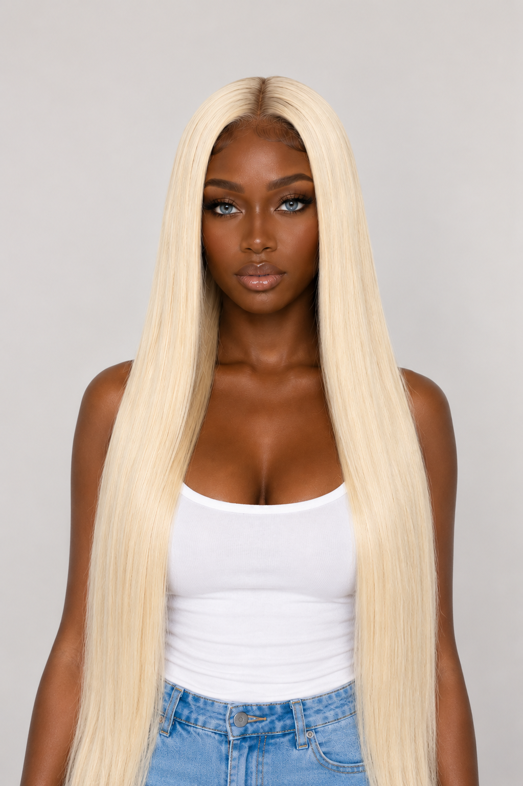 A woman with long, straight, platinum blonde hair extensions wearing a white tank top and blue jeans, photographed against a solid light gray background.