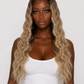 A woman modeling a long, wavy hair extension or wig with a dark brown root and blonde ombre ends, styled in voluminous curls, paired with a white tank top and light blue jeans against a plain light gray background product